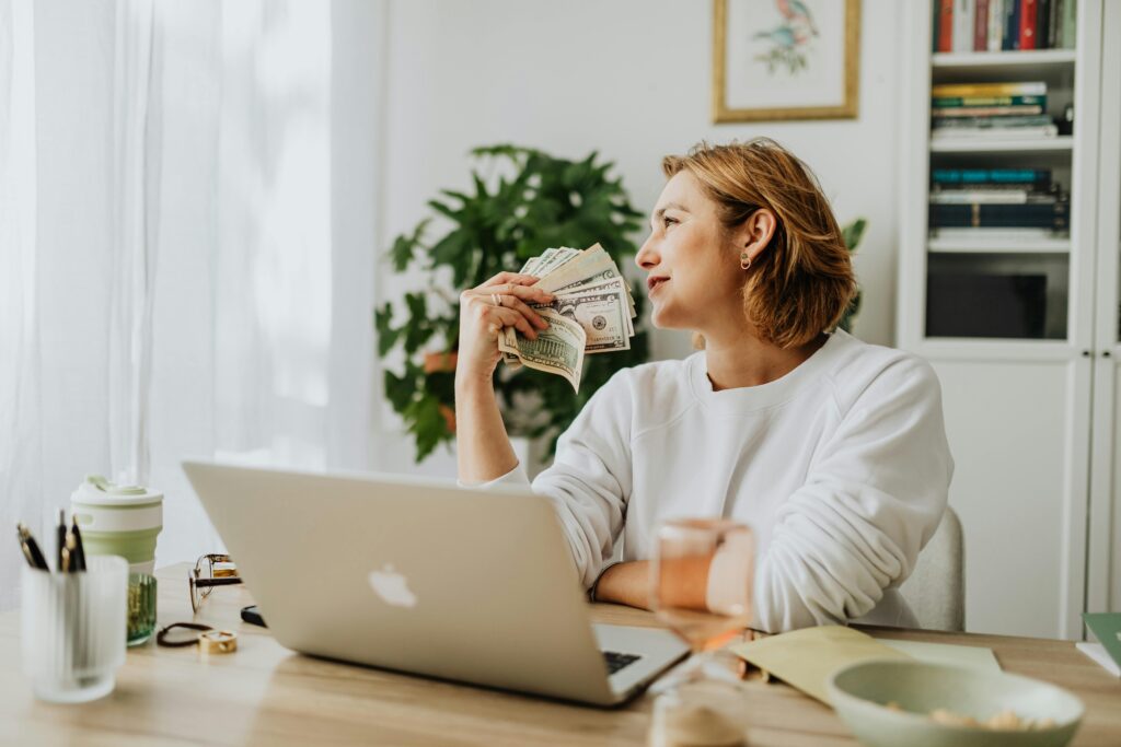 Woman sitting at desk holding cash resources while working on a laptop in a cozy home office setting.